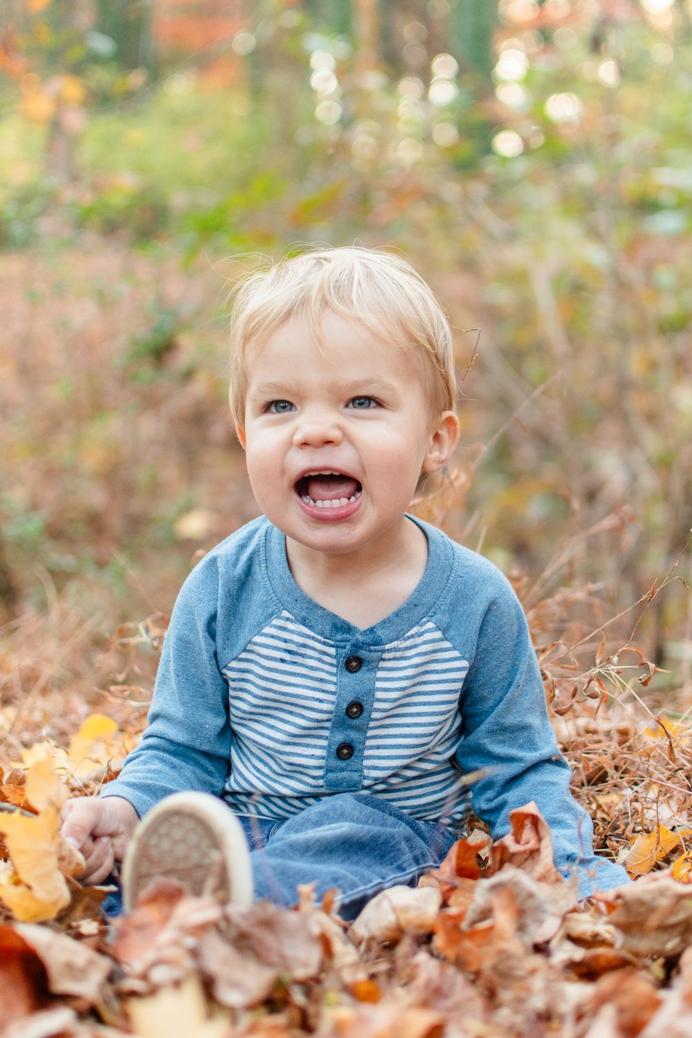 A happy toddler sits among fallen autumn leaves wearing a blue striped shirt.