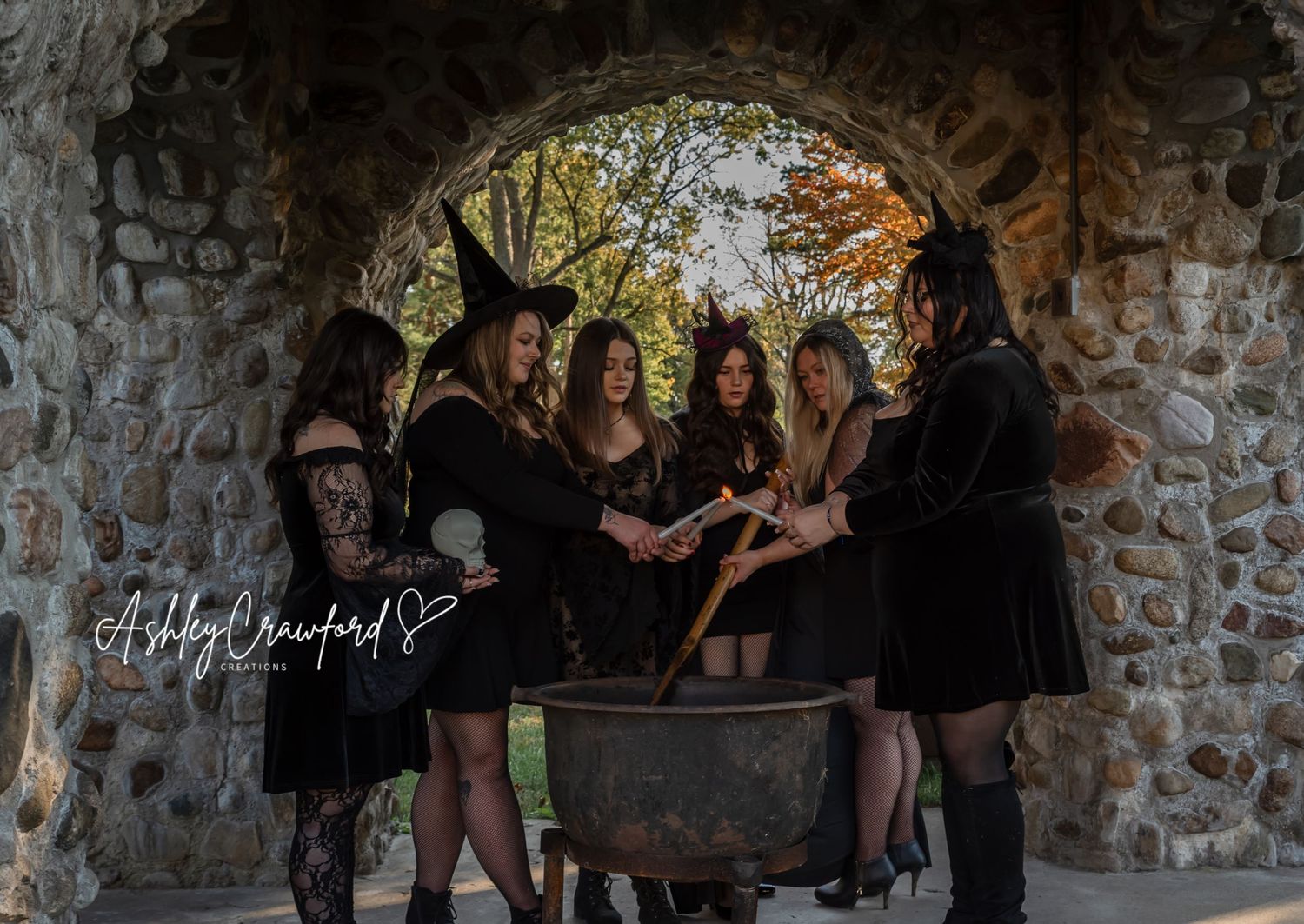 Group in dark attire gathers around a cauldron under a stone archway for a witchy photoshoot.