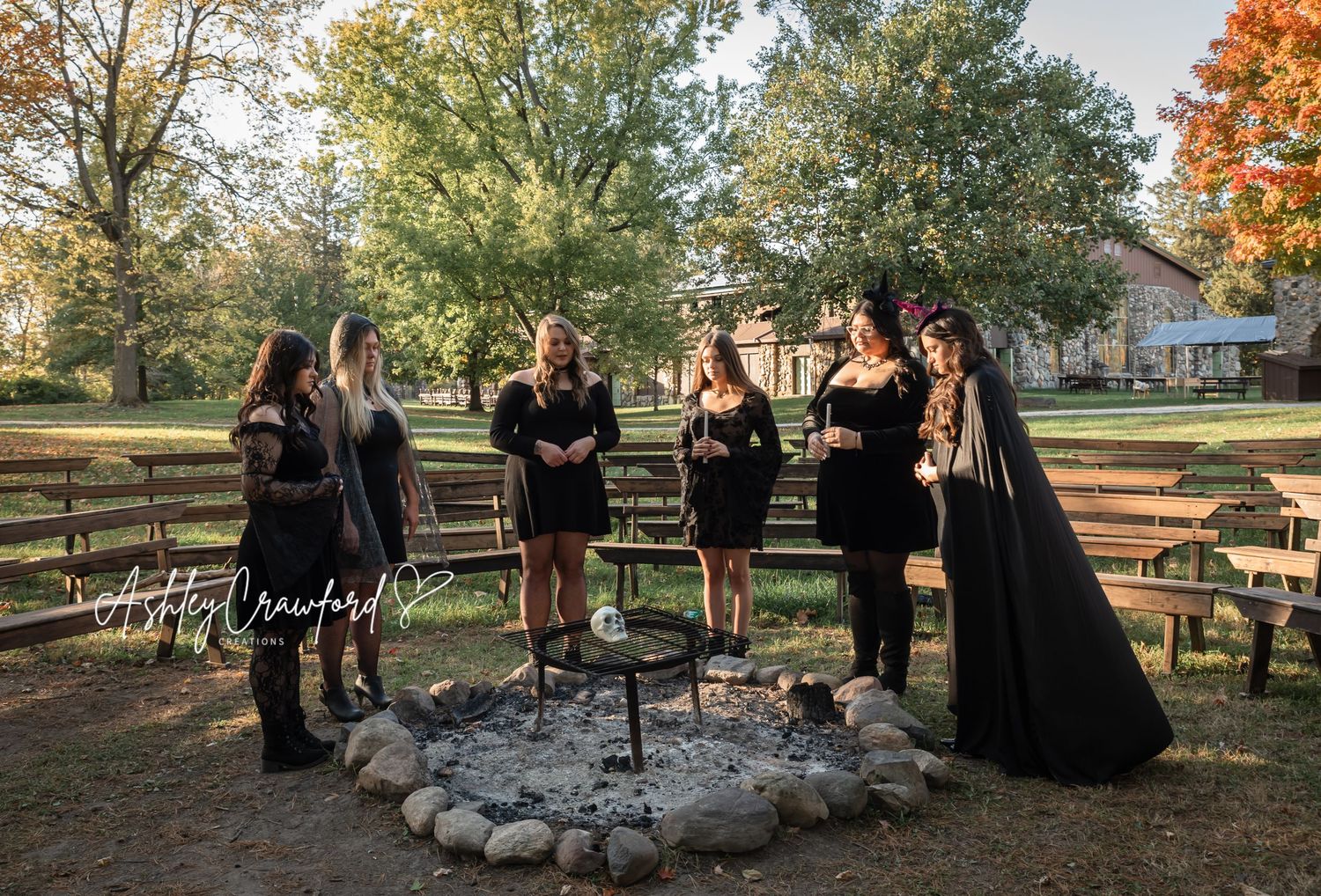 Group dressed in black gathered around a stone fire pit in an autumn outdoor setting.