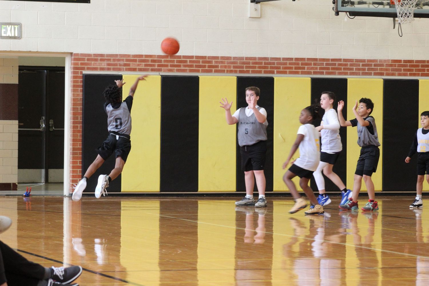 Players on a basketball court during a game with one jumping to make a shot against yellow and black striped wall.