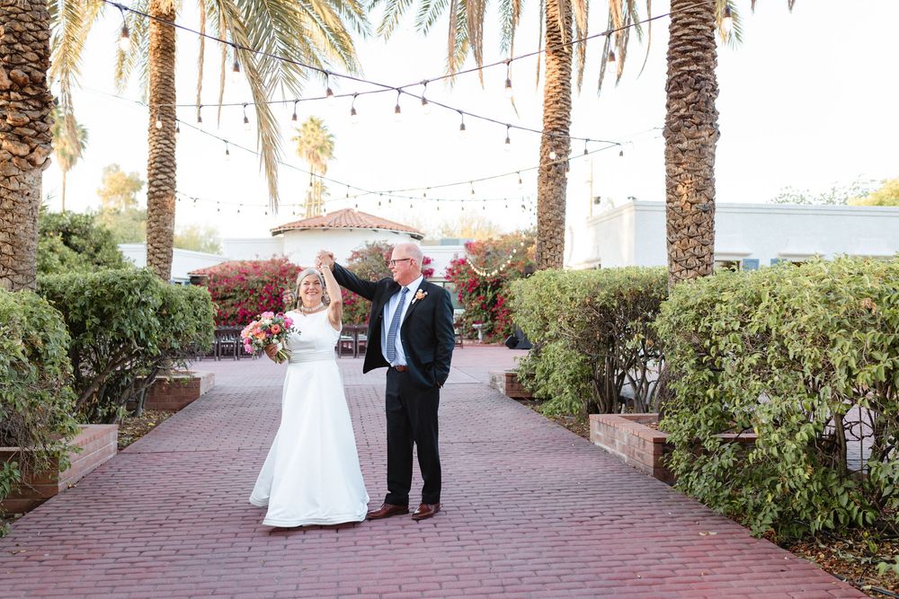A couple stands on a brick path lined with palm trees and string lights at an outdoor venue near the water.