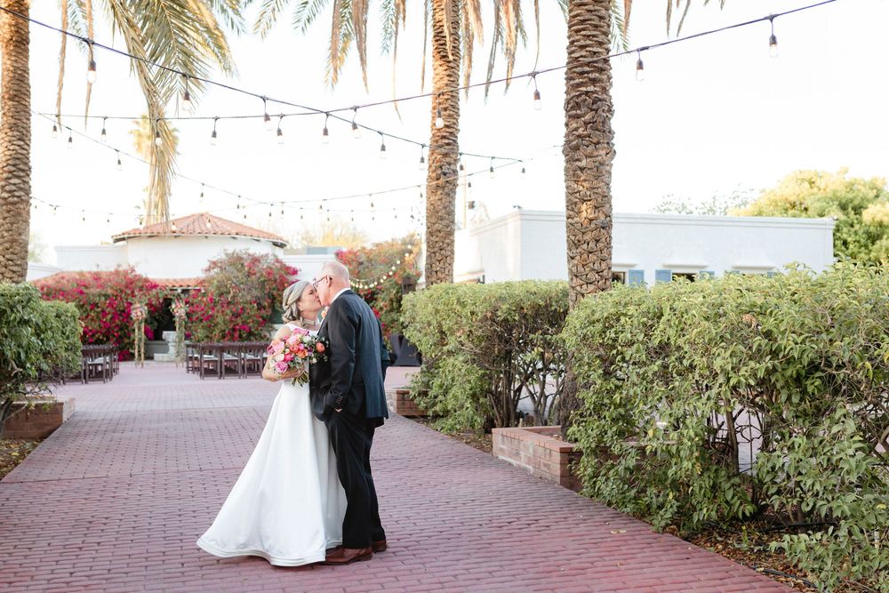 A couple embraces on a brick pathway lined with palm trees and string lights on a sunny day.