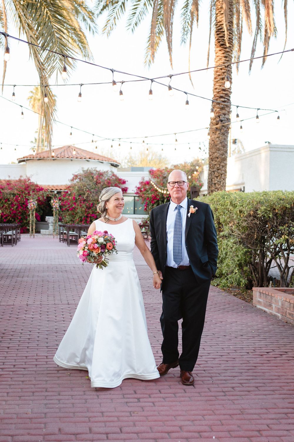 A wedding scene on a brick path under string lights and palm trees with the bride holding a pink floral bouquet.