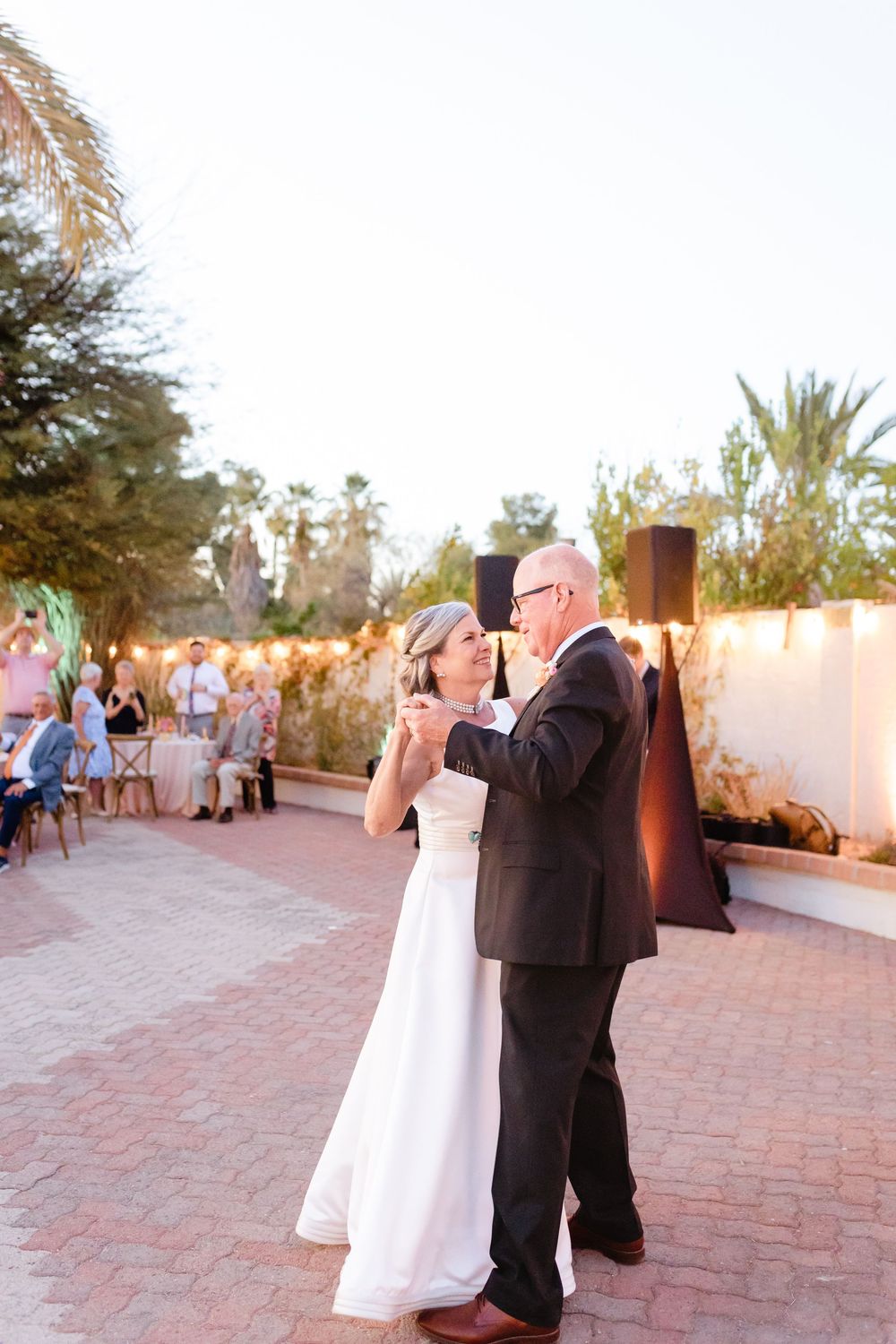 Couple shares a romantic first dance at outdoor wedding reception on brick patio at sunset with palm trees in background.