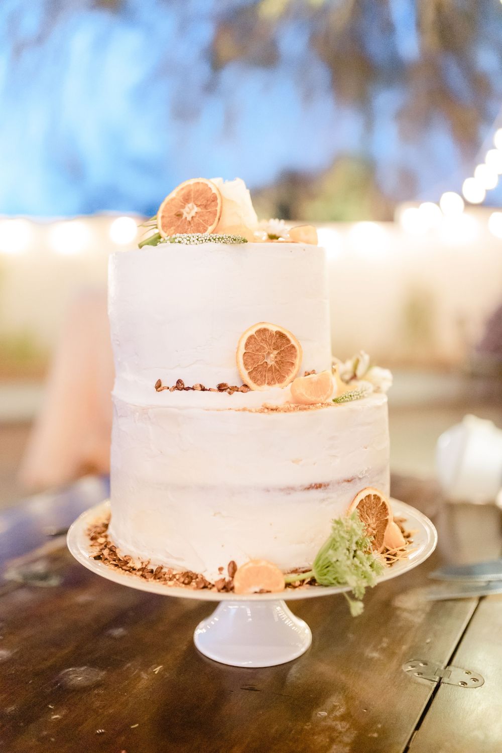 A two-tiered white wedding cake decorated with dried citrus slices and crumbs on a white cake stand.