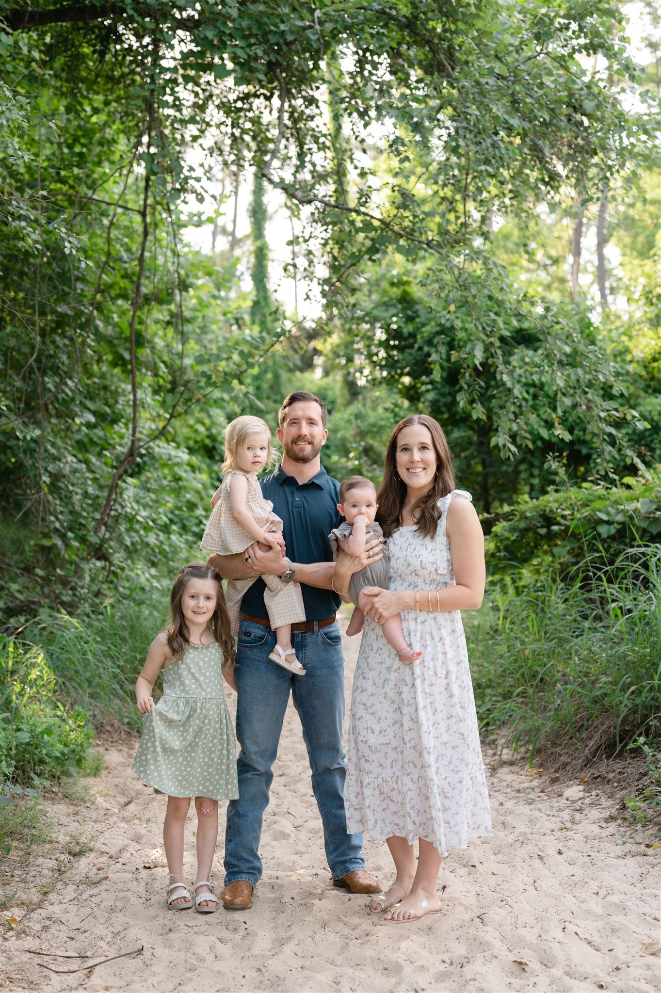 Family stands together on a wooded dirt path surrounded by lush green foliage on a bright summer day.