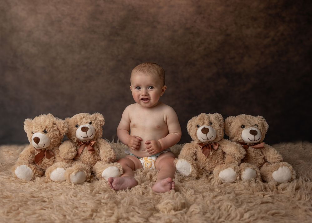 Studio portrait of a small child seated with a collection of brown teddy bears on a textured beige background.