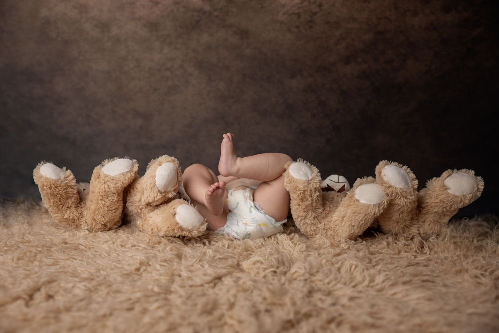 A baby lies on a soft fur rug wearing a white diaper with teddy bears arranged around them in a cozy studio setting.