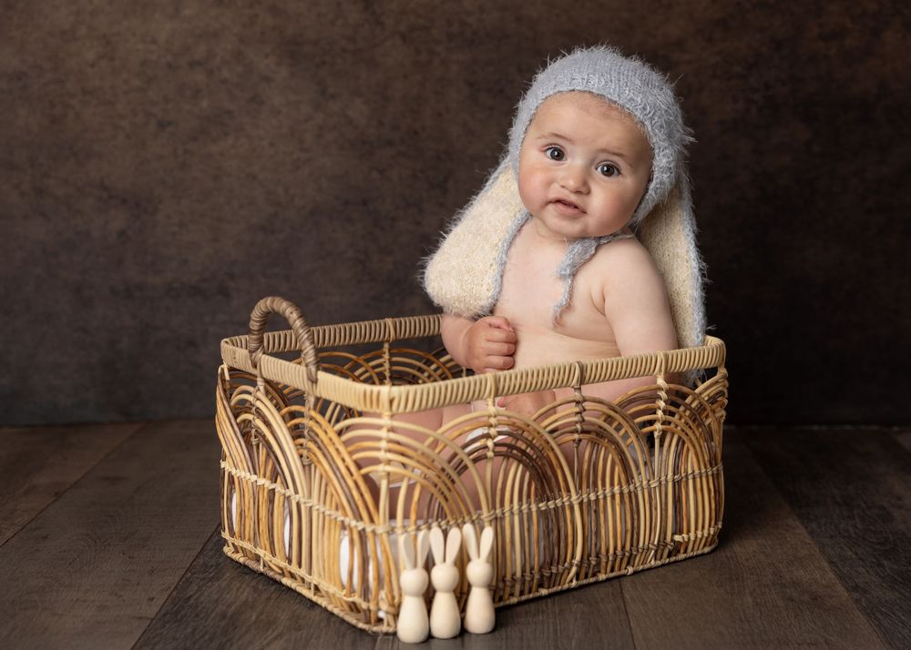 A baby wearing a white knit hat sits in a woven wicker basket against a dark brown backdrop.