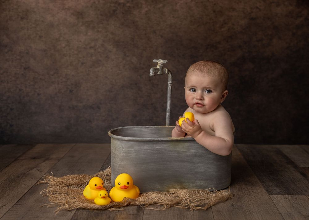 A baby sits in a metal wash tub with rubber duckies on a wooden floor against a dark textured background.