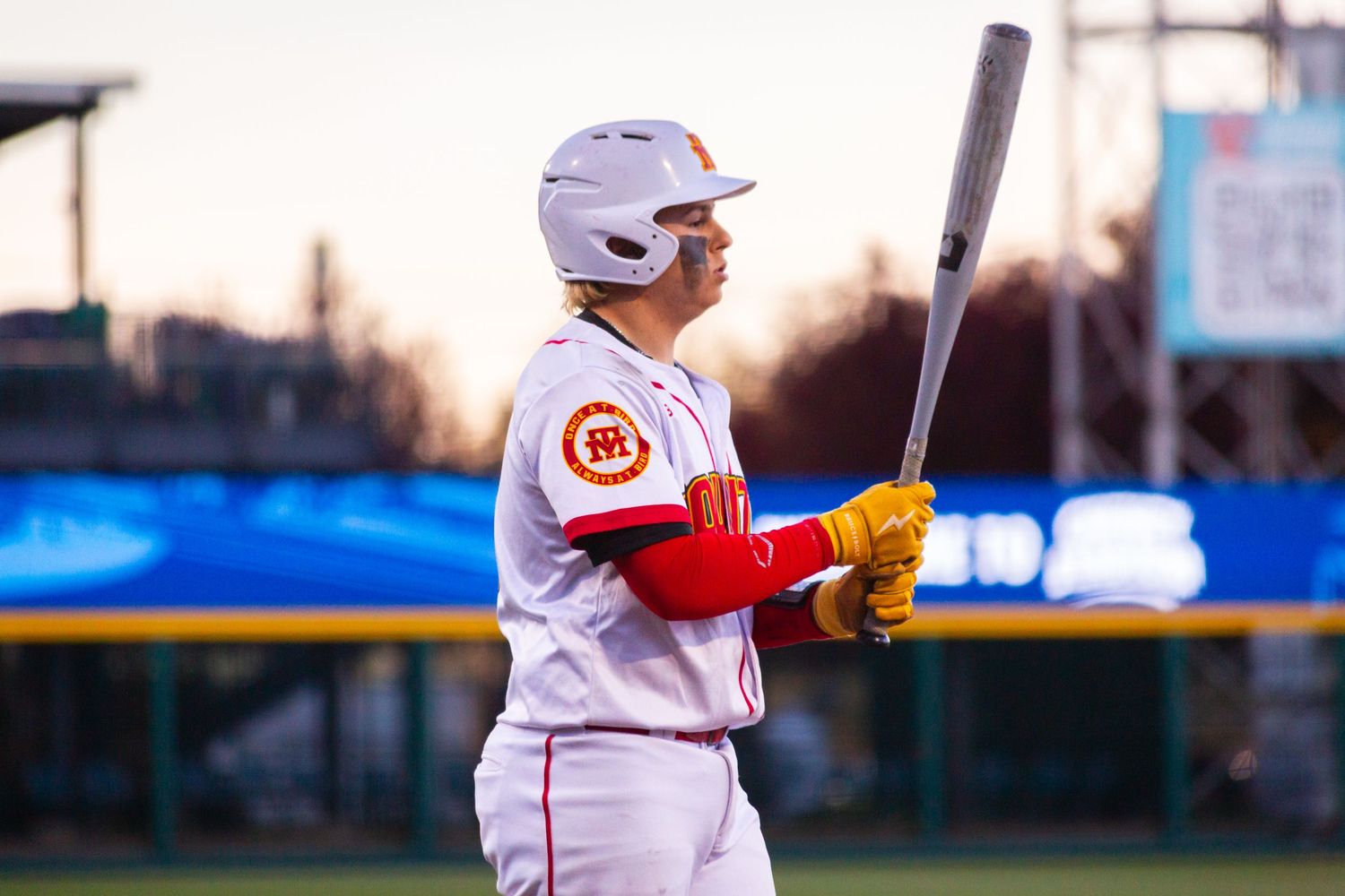 A baseball player in a white and red uniform stands at bat during evening practice, holding their stance at twilight.
