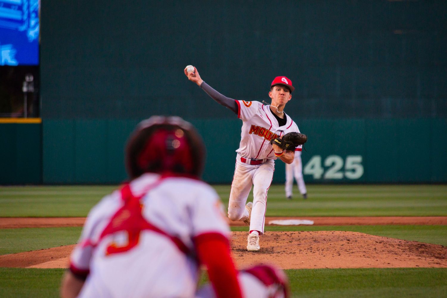 Baseball pitcher in white uniform throws a pitch from the mound as batter in red waits at home plate during night game.