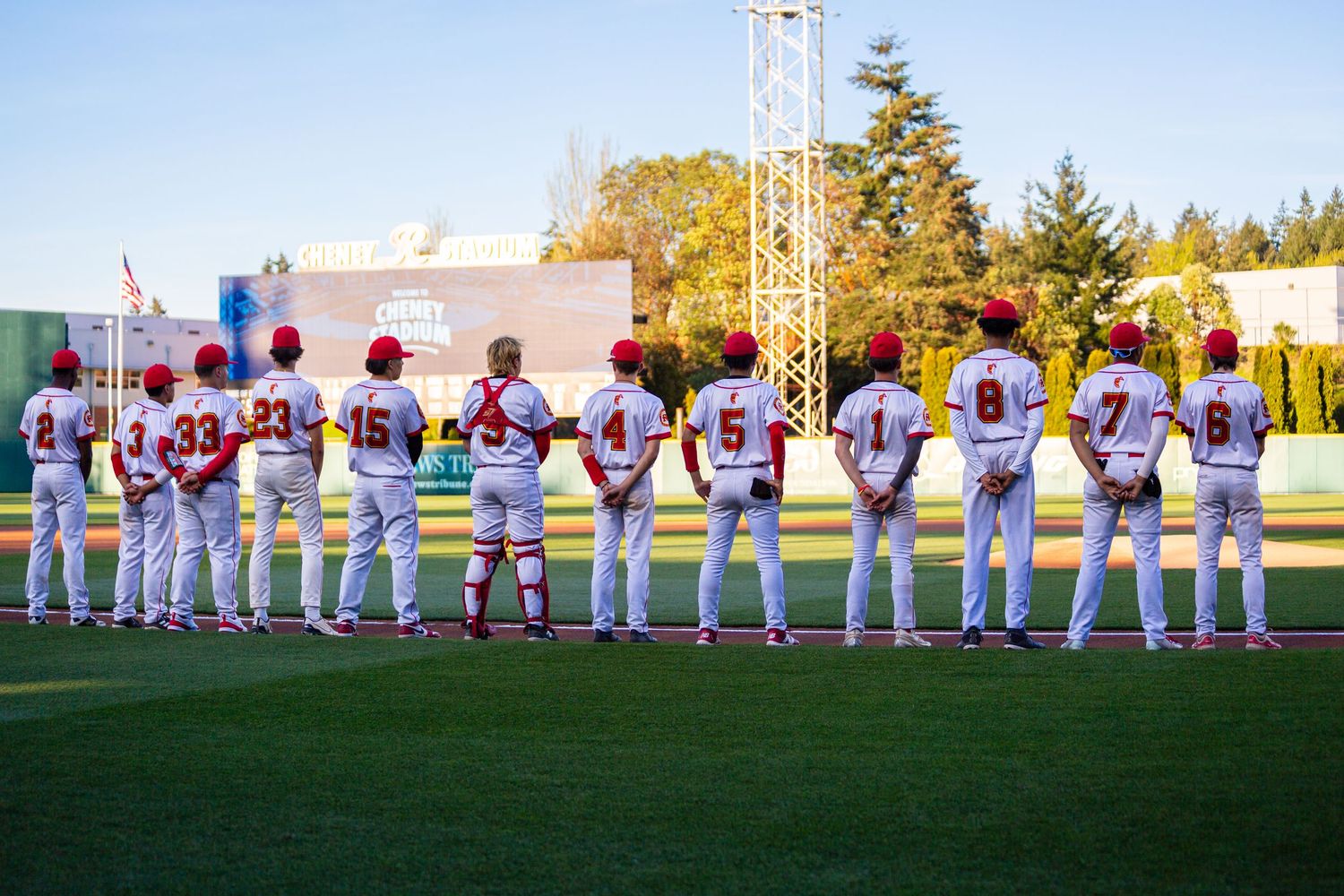Baseball team lines up along the first base line wearing white uniforms during pregame ceremonies on a sunny evening.