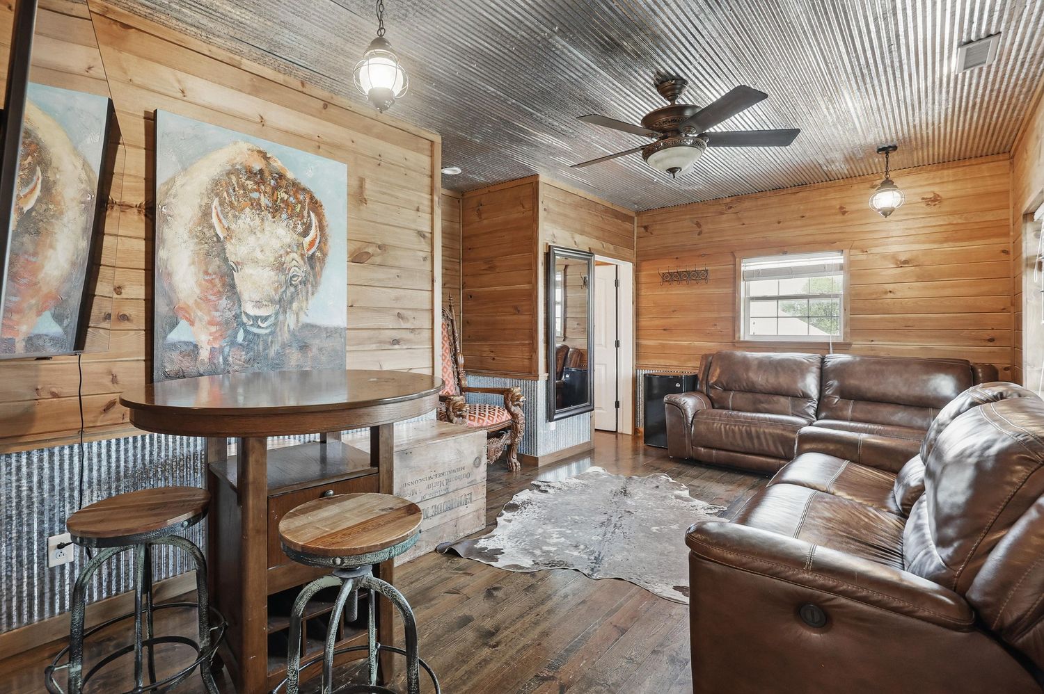 Rustic cabin interior with leather sectional sofa, wood walls, corrugated metal ceiling, and wildlife artwork.