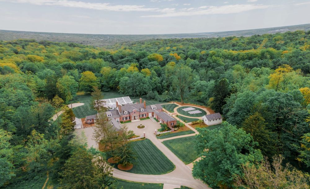 Aerial view of a grand estate with manicured lawns surrounded by lush forest landscape.