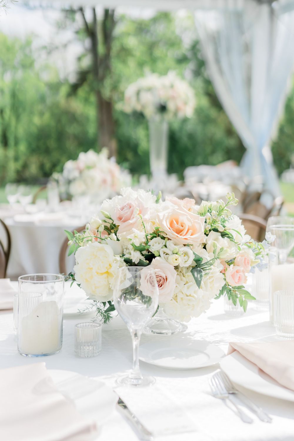 Elegant wedding table centerpiece with blush roses and white flowers in a crystal vase on white linen tablecloth.