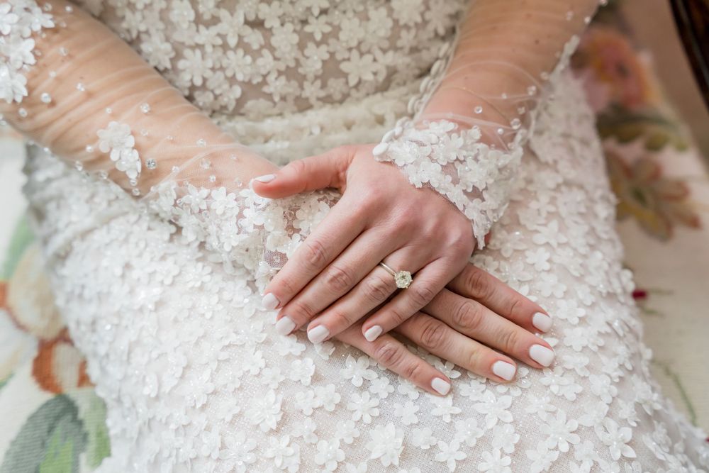 Close-up of bride's hands showing elegant diamond ring and intricate lace sleeve details.