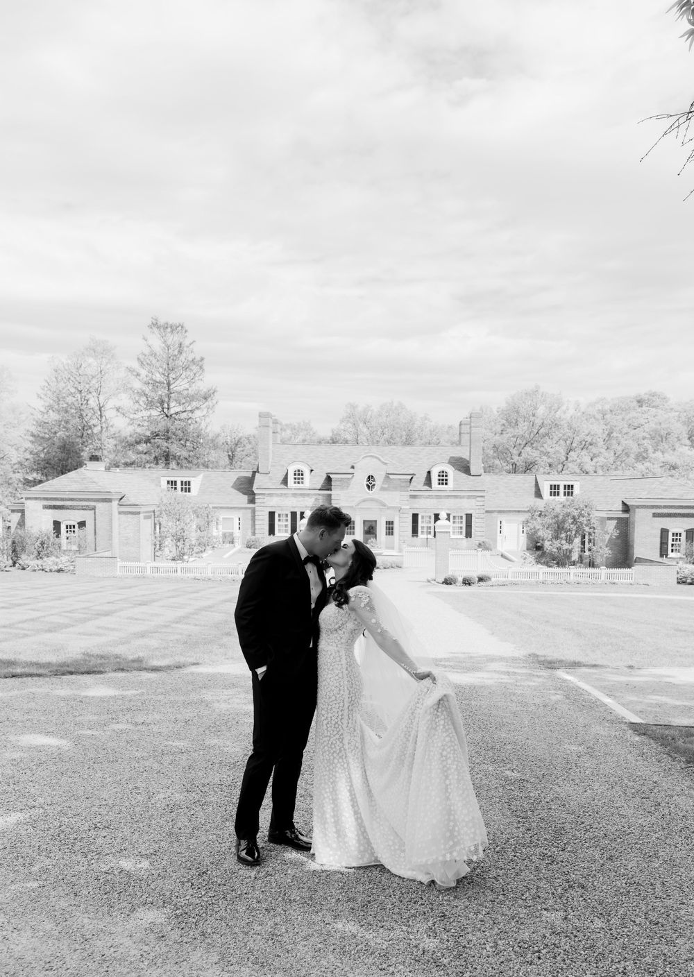 A couple in formal wedding attire share an intimate moment on the lawn of an elegant estate with dormered windows.