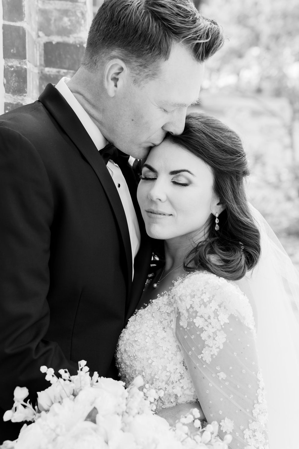 Black and white portrait of newlyweds sharing an intimate moment in formal wedding attire against brick wall background.