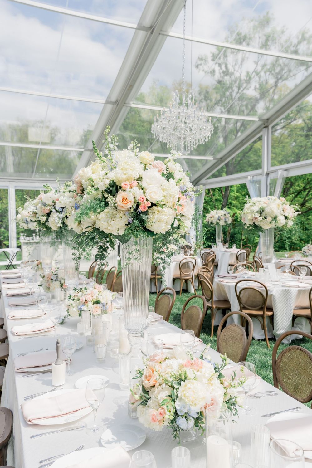 Elegant wedding reception tables set in clear tent with peach and white floral centerpieces and crystal chandelier overhead.