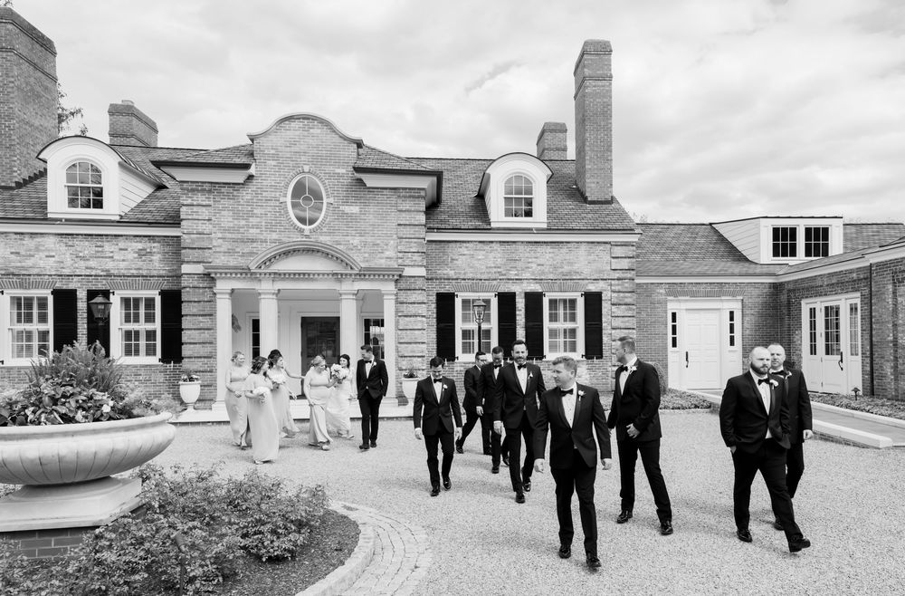 A formal gathering of people in dark suits walking on a curved driveway outside an elegant colonial-style mansion.