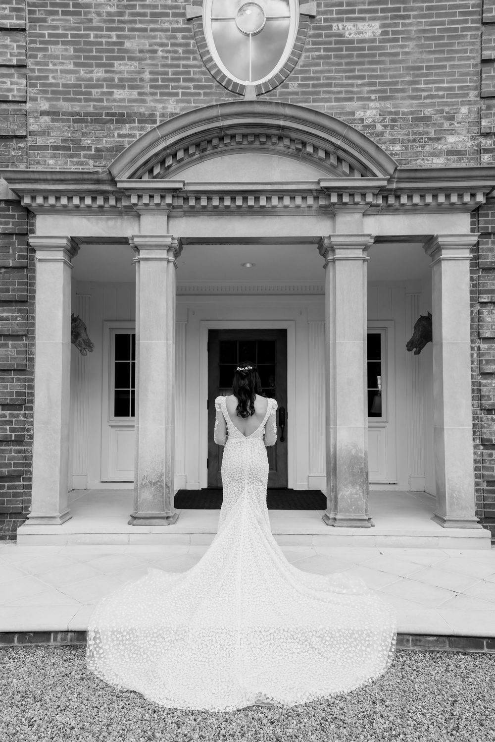 A stunning lace mermaid wedding gown with a long train photographed from behind in front of an elegant brick entrance.