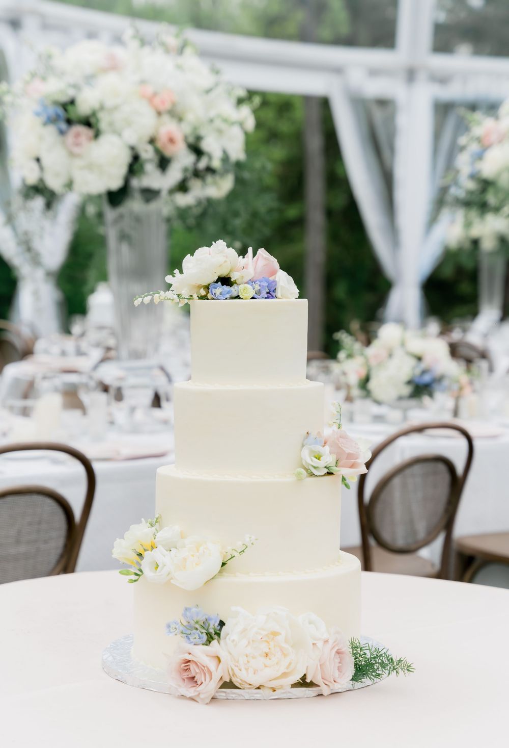 Elegant four-tier white wedding cake decorated with blush roses and blue accents on display at outdoor reception venue.