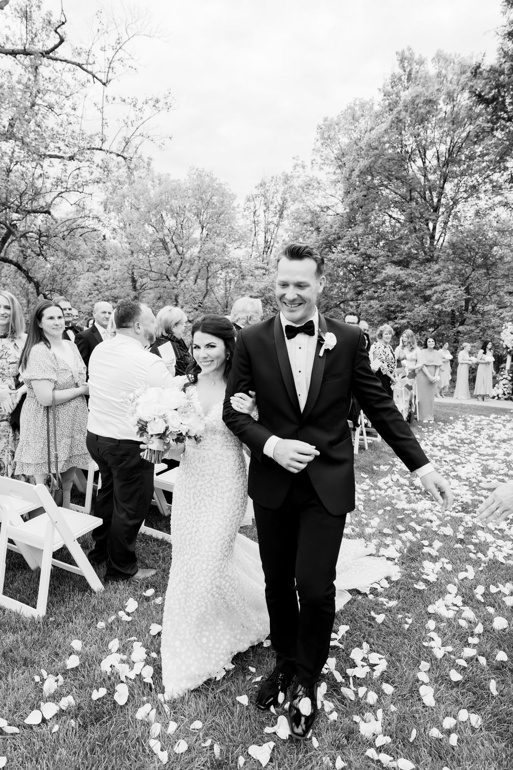 A couple walks joyfully down an outdoor wedding aisle covered in flower petals while guests look on.