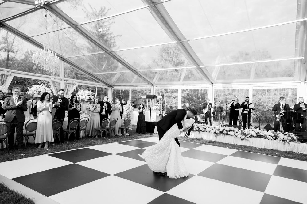 A couple shares their first dance on a black and white checkered dance floor under a clear tent at their wedding.