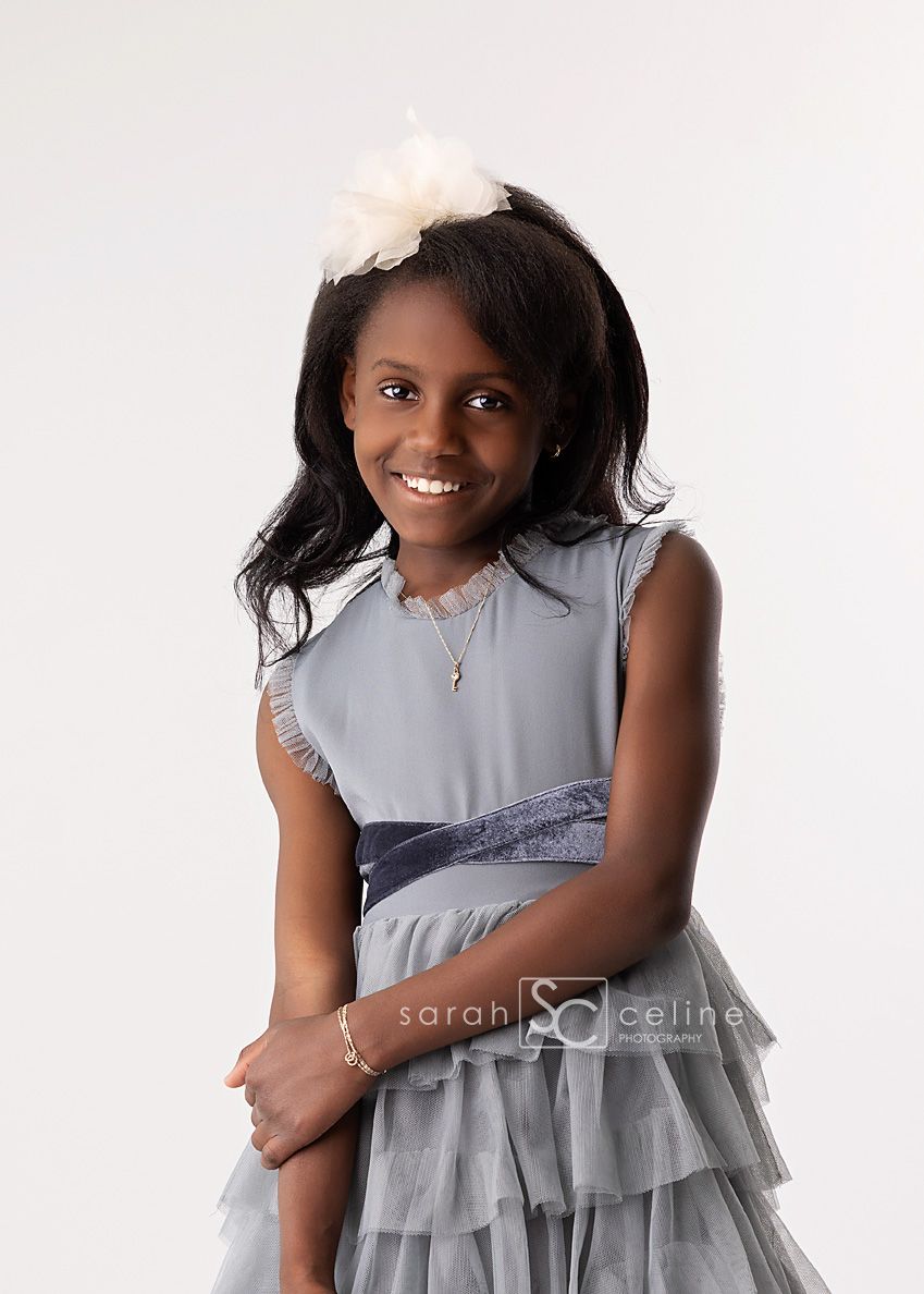Studio portrait of a person in a light gray sleeveless dress with ruffled layers and a white floral hair accessory.