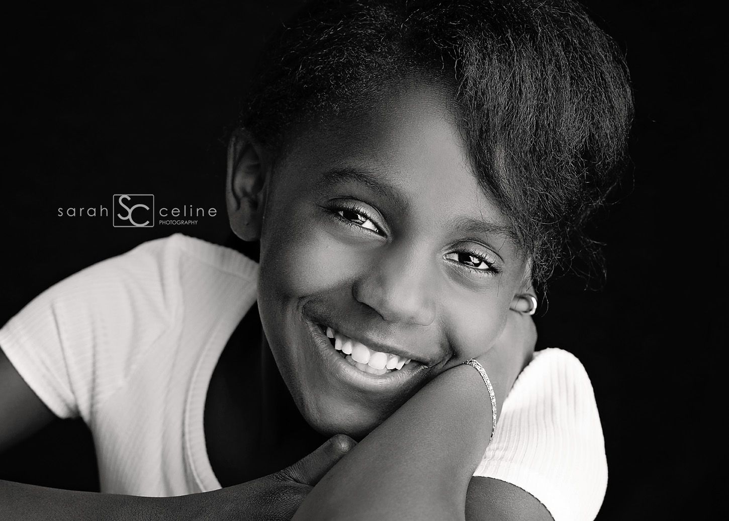 A black and white portrait showing a warm, radiant smile and expressive eyes while resting on hands in a casual pose.
