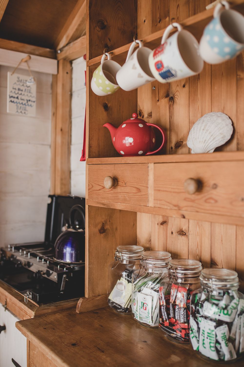 Cozy kitchen corner with hanging teacups, red teapot, glass storage jars and wooden cabinets in rustic cottage style.