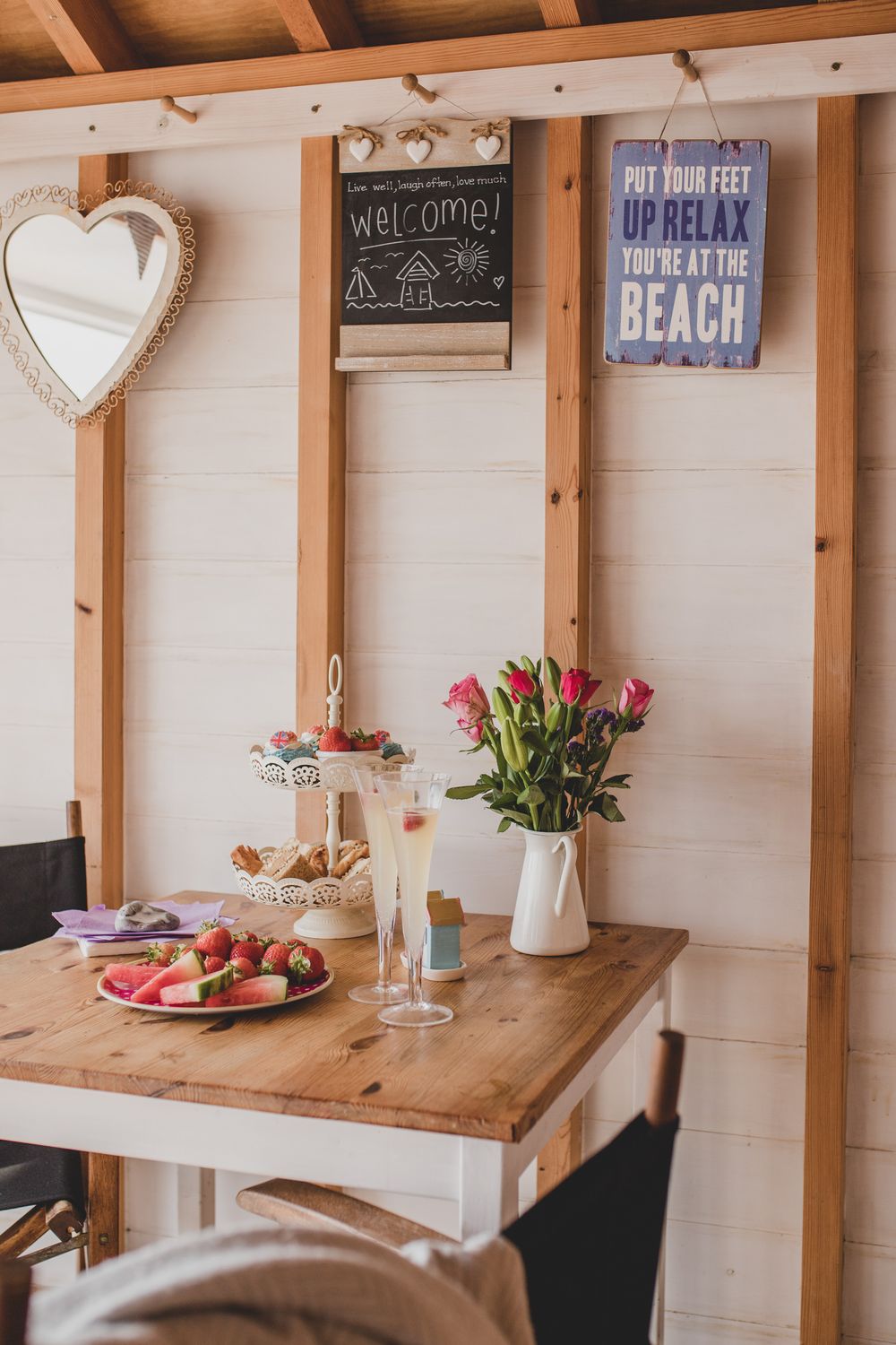 Cozy beach-themed dining nook with wooden table, pink flowers, fresh fruit plate and welcome signs on white paneled wall.