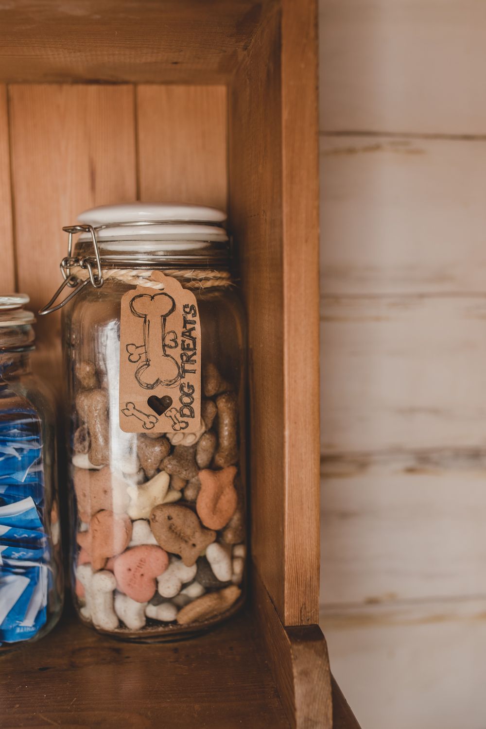 A glass jar filled with dog treats sits on a wooden shelf, with a handwritten label visible through the clear container.