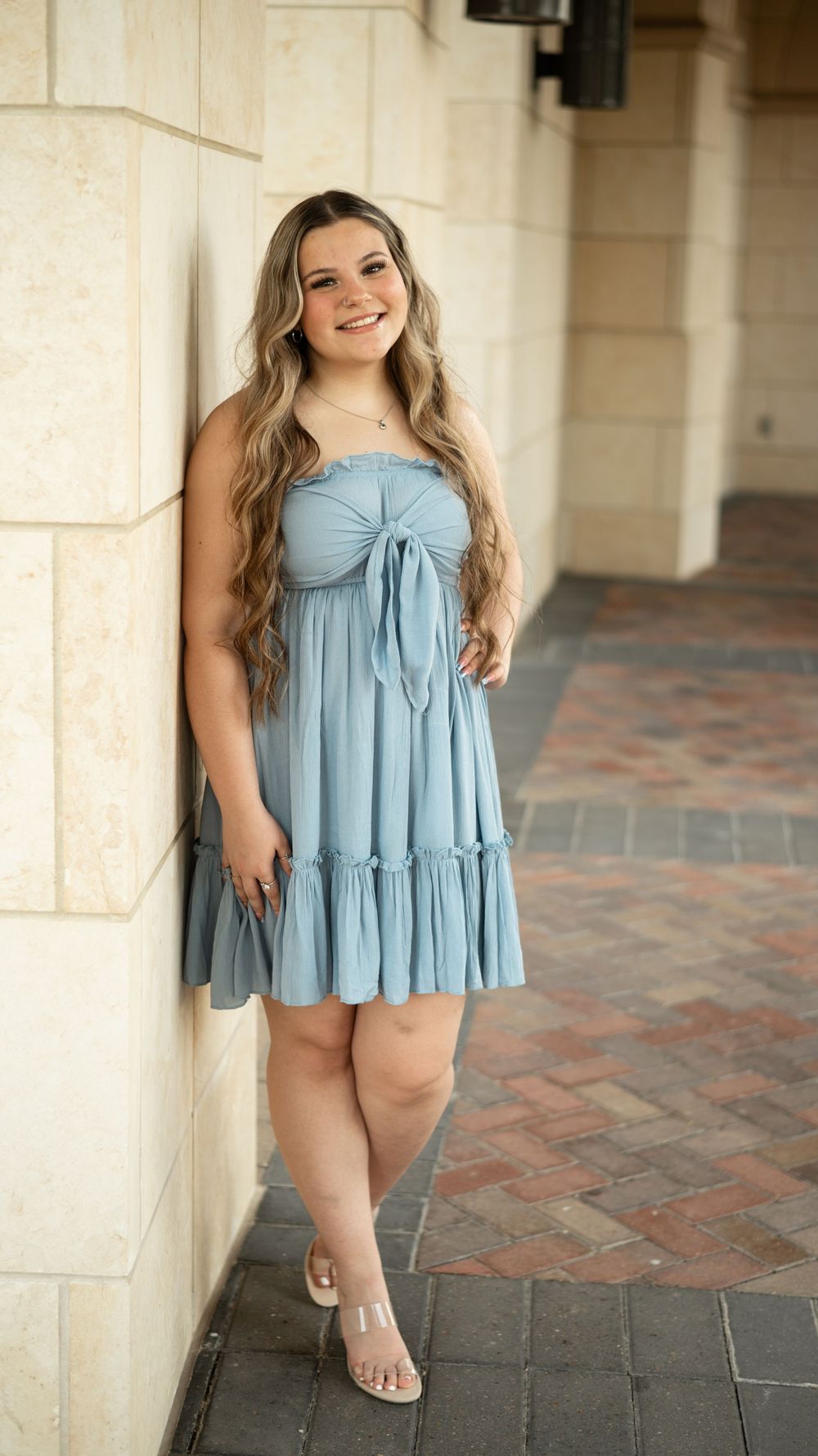 Someone wearing a light blue summer dress with tiered ruffles poses against a beige stone wall on brick pavement.