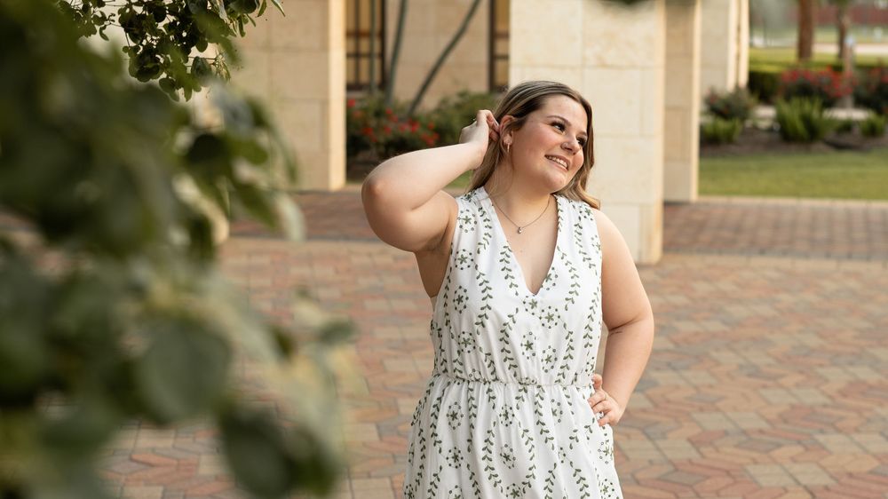 A person in a white floral dress poses on a brick pathway surrounded by greenery on a sunny day.