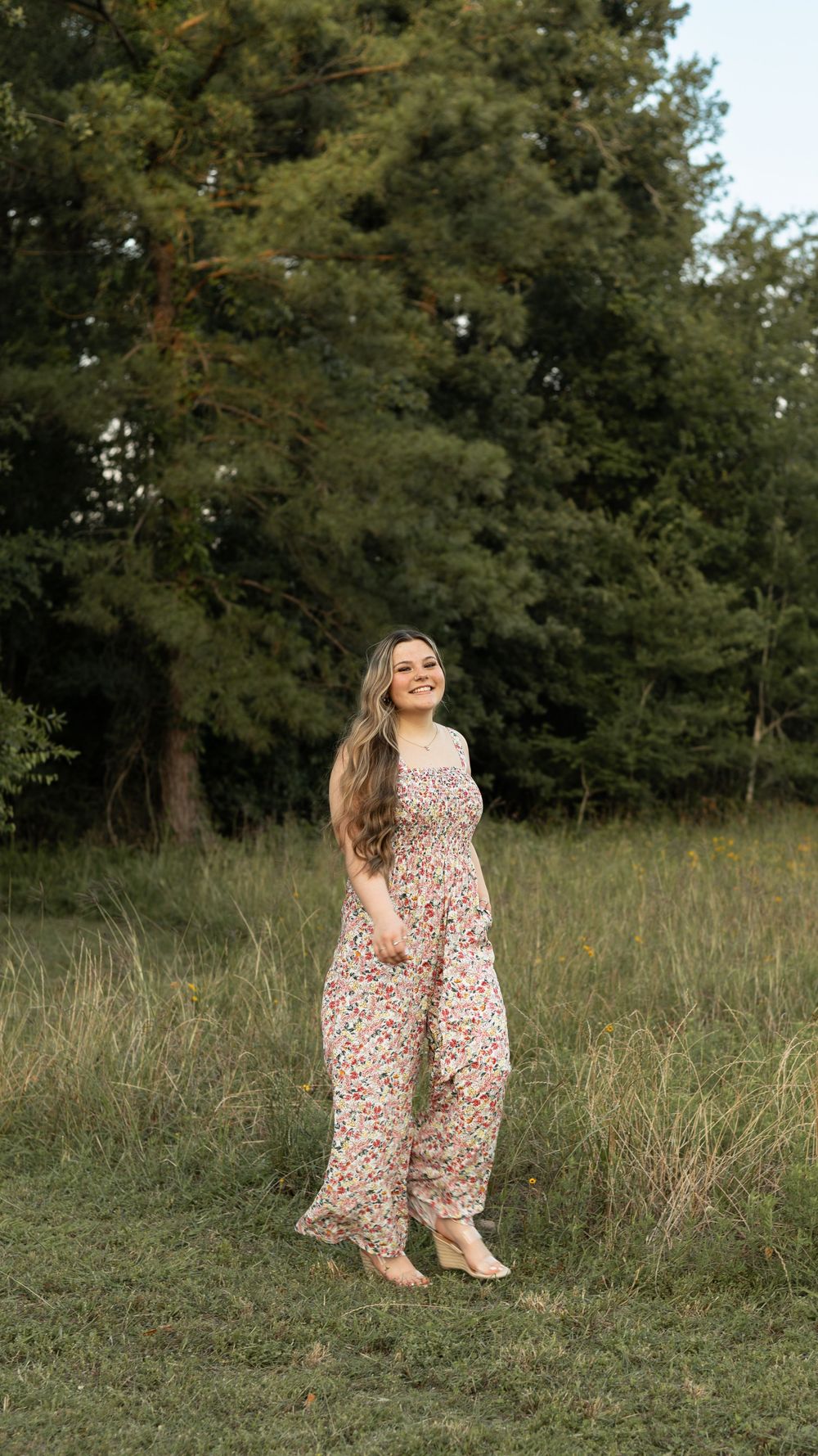 A person in a floral maxi dress stands in a grassy field with pine trees in the background.