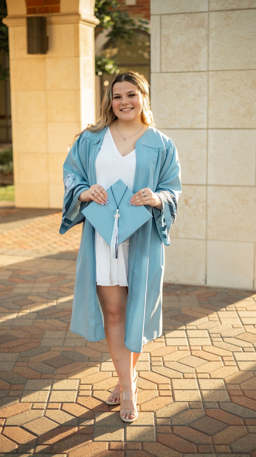Graduating student wearing light blue graduation gown and white dress poses on brick pathway during golden hour celebration.