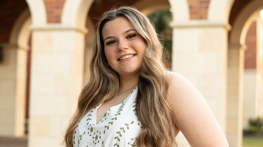 Smiling young woman stands in front of outdoor arches wearing a white sleeveless floral dress and long blonde hair.