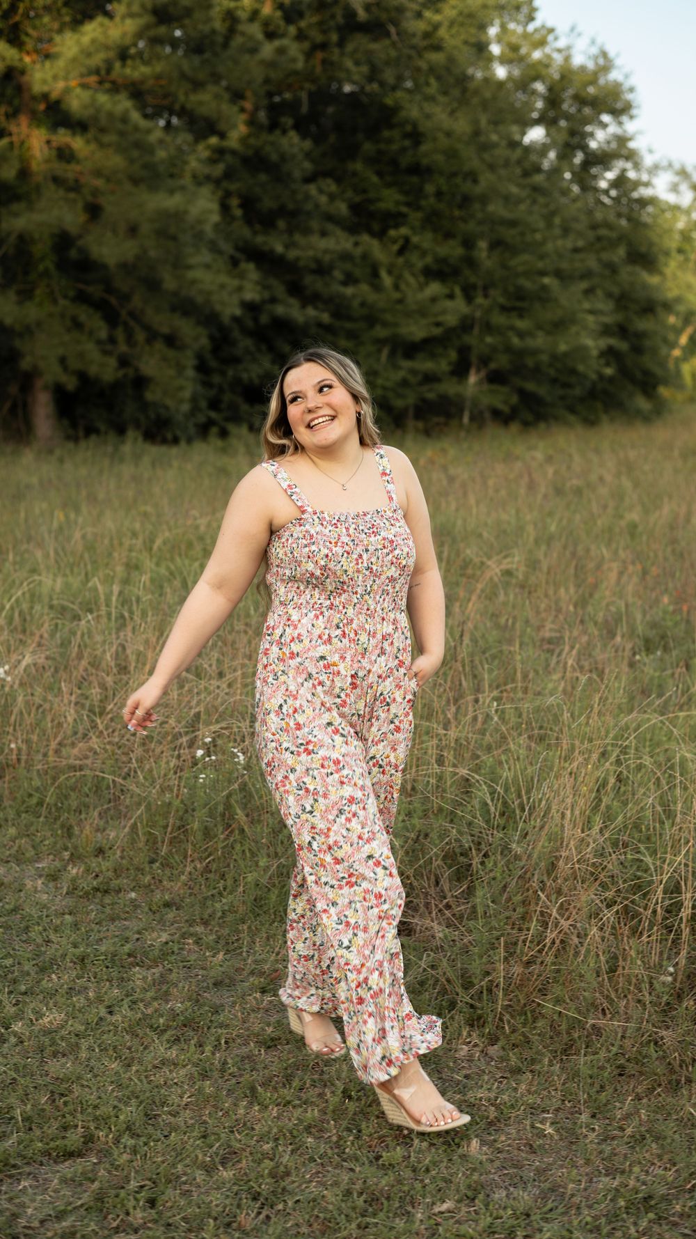 A person wearing a floral maxi dress poses joyfully in a grassy field surrounded by trees on a summer evening.