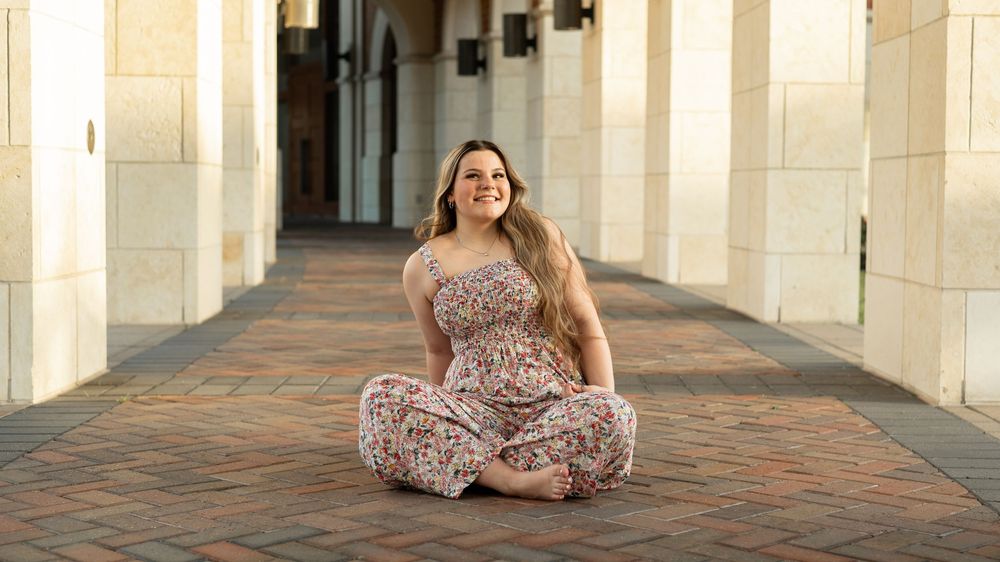 A person in a floral jumpsuit sits cross-legged on a brick walkway framed by columns.