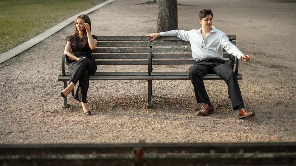 Two people sit at opposite ends of a park bench on a gravel path, displaying emotional distance between them.