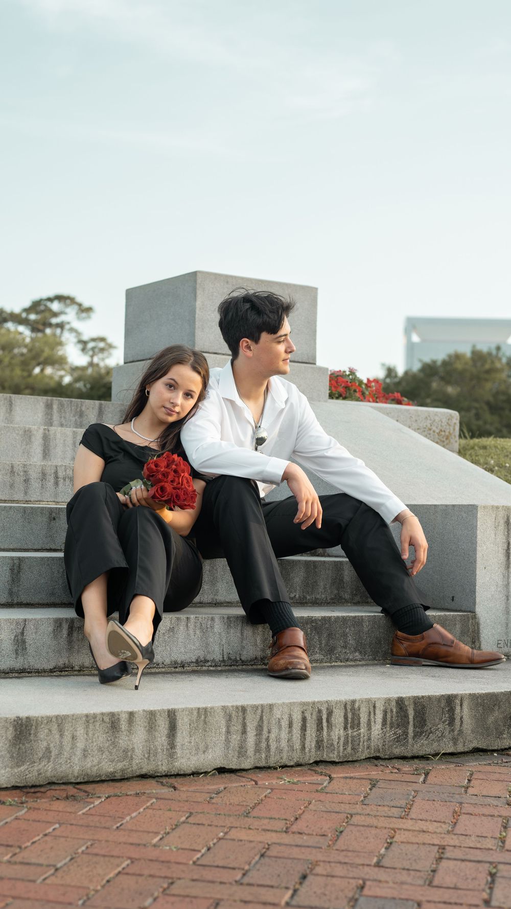A couple in contrasting black and white outfits sit together on concrete steps with red flowers near modern architecture.