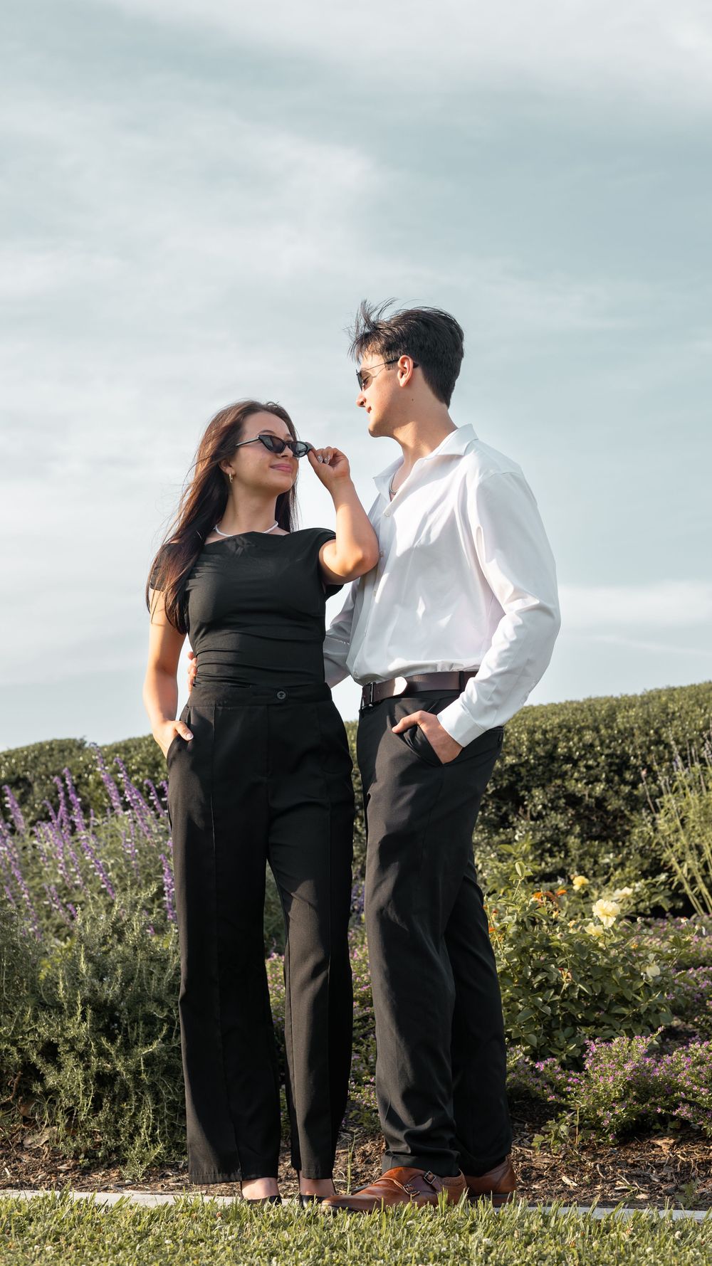 A couple stands together outdoors in formal attire amid blooming wildflowers and a cloudy sky.