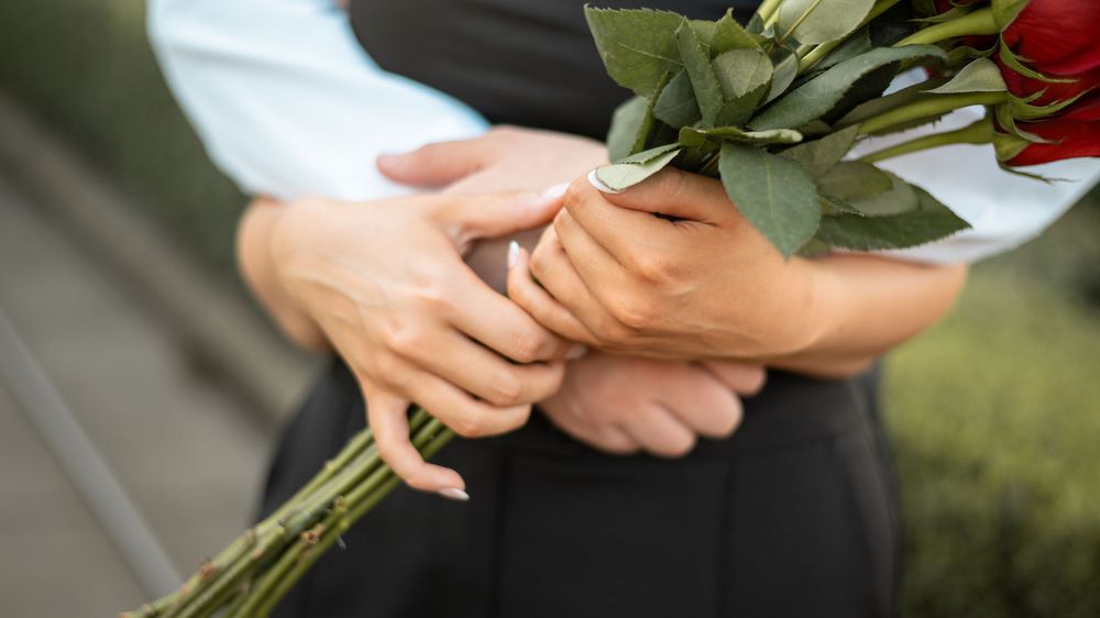 Hands holding a bouquet of red roses with green leaves against a black dress in soft natural lighting.