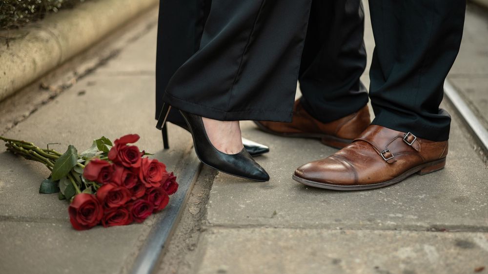 Two pairs of feet in dress shoes stand on concrete next to a bouquet of red roses.