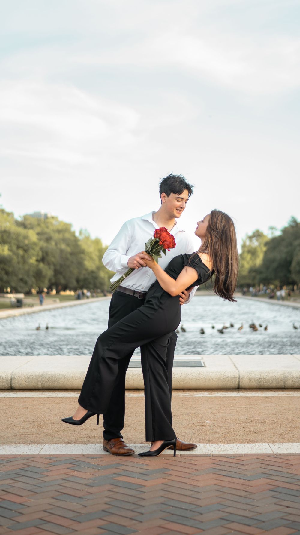 A romantic couple dances by a fountain holding red roses while dressed in formal attire against a scenic outdoor backdrop.