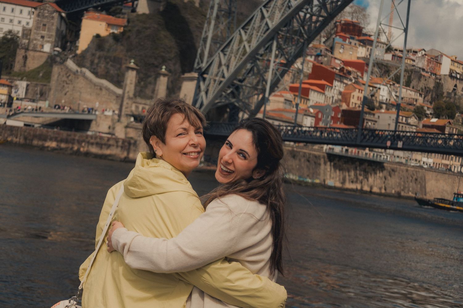 Two friends hugging and laughing by the riverside with Porto's iconic bridge in the background.