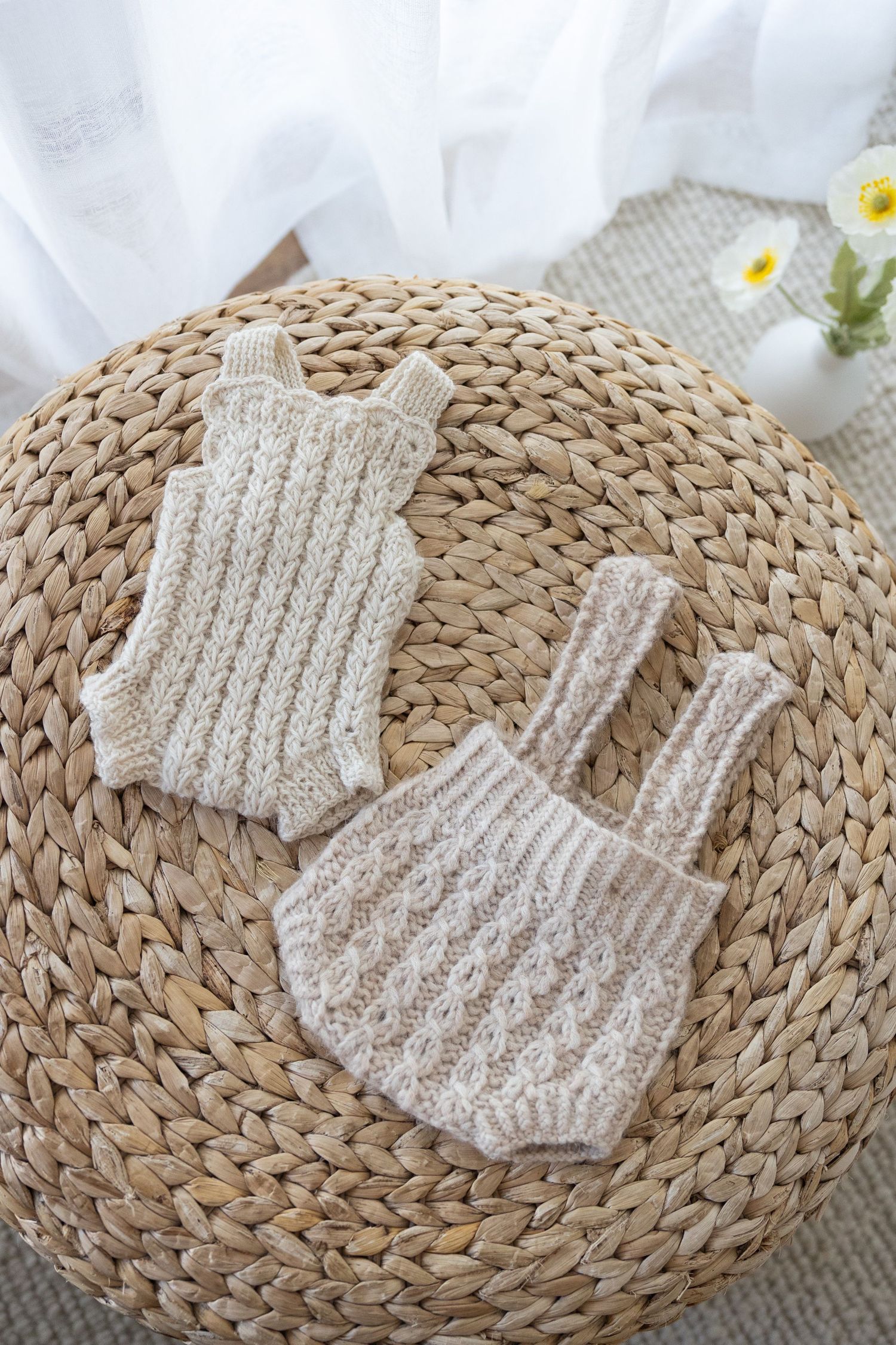 Two handknit beige baby rompers displayed on a natural woven straw placemat with white fabric and daisies in background.