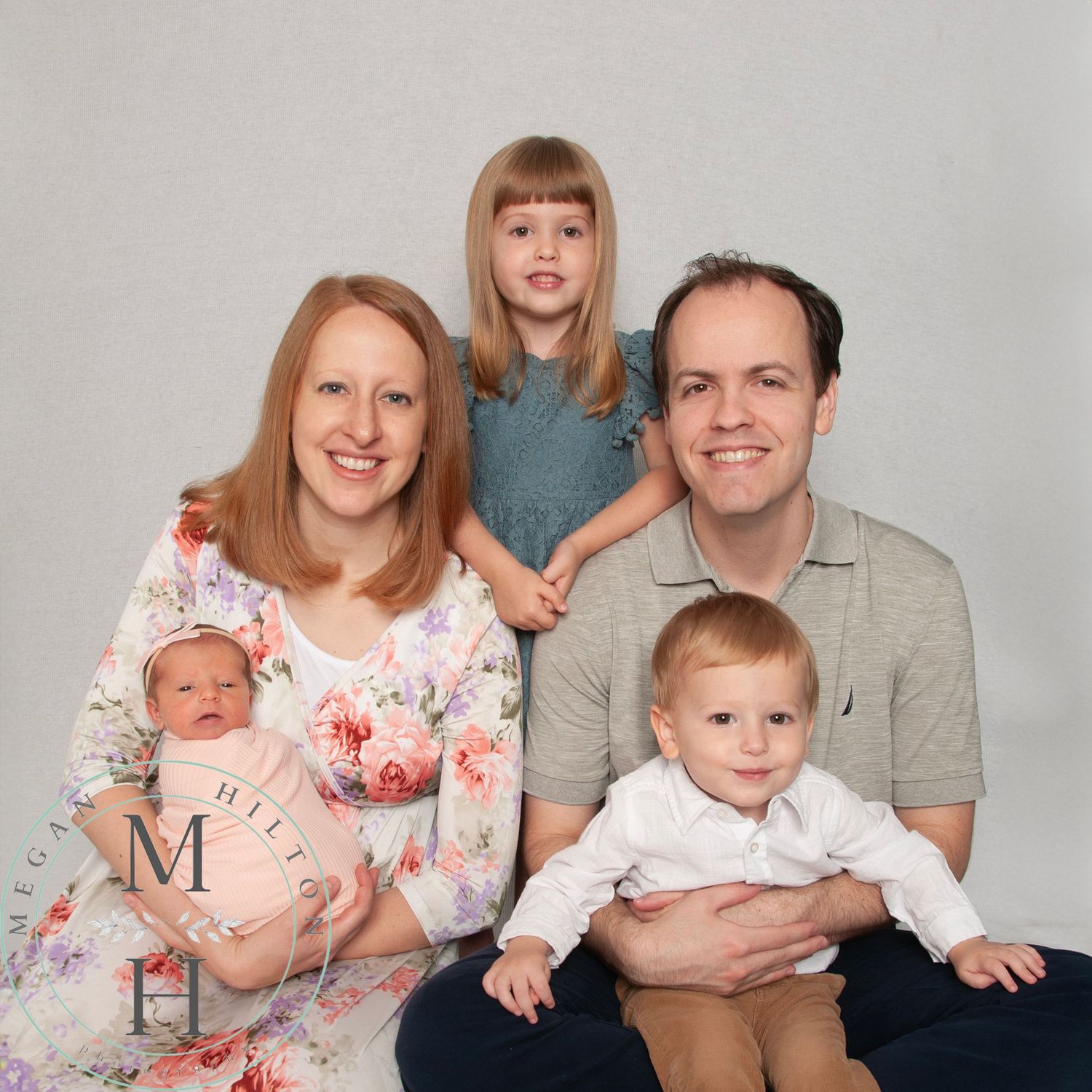 Studio-style portrait of a young family with parents and three children sitting together against a neutral background.